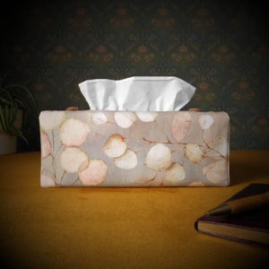 May include: A tissue box cover with a floral design featuring delicate, light pink and beige leaves on a grey background. The cover has a rectangular shape and is made of fabric.