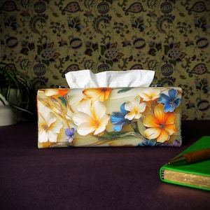 May include: A tissue box cover with a floral design. The cover features white, orange, yellow, blue, and green flowers on a cream background.