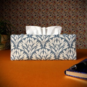 May include: A rectangular tissue box cover with a blue and white leaf pattern. White tissues protrude from the top. The box is on an orange surface, with a notebook and pen in the background.
