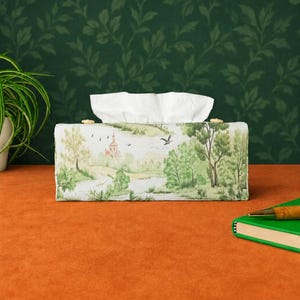 May include: A rectangular tissue box cover featuring a printed landscape scene with green trees, a small building, and birds in flight. The fabric cover has a light background and is topped with white tissues. Two small wooden buttons are visible on the top.