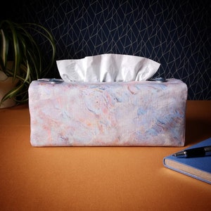 May include: A rectangular tissue box cover with a pastel marbled design in shades of pink, blue, and orange. White tissues are visible at the top. The cover sits on a brown surface with a blue notebook and pen.