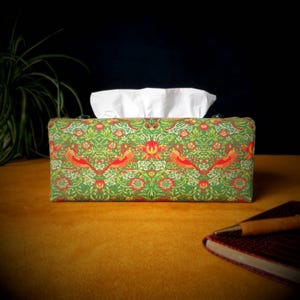 May include: A green tissue box cover with a floral pattern and red birds. The cover has a white tissue box inside.