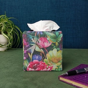 May include: A square tissue box cover featuring a vibrant botanical print with large pink and yellow flowers, green foliage, and a prominent dragonfly. The fabric surface displays a detailed, colourful pattern of various blossoms and leaves.