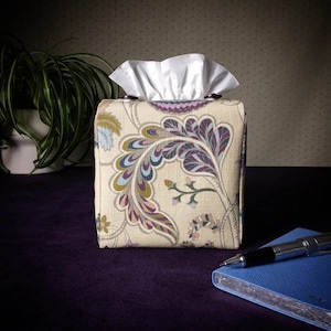 May include: A beige tissue box cover with a floral pattern in shades of purple, blue, and green. The cover has two brown buttons on the top.