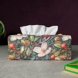 May include: Rectangular tissue box with a floral pattern of white magnolia flowers, green leaves, and red buds on a dark teal background. The box has gold-coloured accents and white tissues are visible.