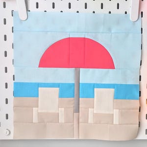Peut inclure: Un bloc de courtepointe représentant une scène de plage avec deux chaises blanches sous un parasol rouge sur une mer bleue et du sable beige.