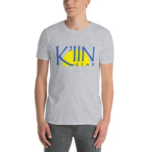 May include: A light gray t-shirt featuring the text "K'IIN GEAR" in blue, with a yellow sun design. The shirt is made of a soft material and is suitable for casual wear.