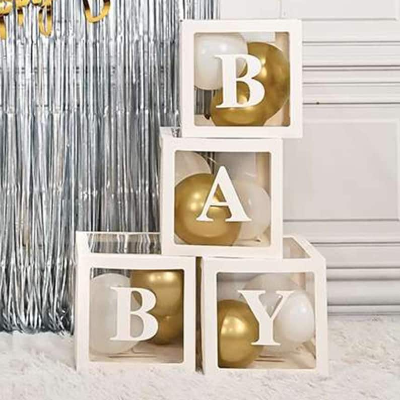 May include: Four white cube boxes with clear sides, stacked on top of each other. Each box contains gold and white balloons and a white letter, spelling out the word "BABY".