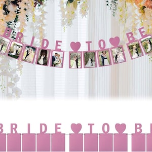Puede incluir: Pancarta rosa de 'Bride to be' con marcos para fotos. La pancarta tiene las palabras 'bride to be' con corazones entre las palabras. La pancarta está colgada de una pared blanca.