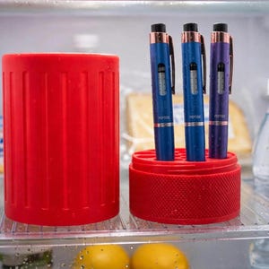 May include: A bright red cylindrical storage container with a matching circular base featuring multiple slots. Three medical-style pens with blue and purple metallic bodies and black caps are inserted into the base, each labelled with the word PEPTIDE.