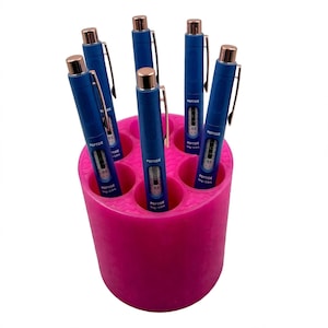 May include: A bright pink cylindrical holder with six circular slots containing blue pens. Each pen features a rose gold top, a silver clip, and a clear window displaying a scale with the text PEPTIDE 5mg + 0.5mL printed on the side.