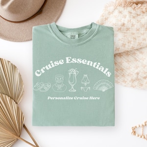 May include: A seafoam green t-shirt with the words "Cruise Essentials" in a white, arched font. Below the text are white line drawings of sandals, a sailor, a cocktail, a bikini, and a fan. The text "Personalize Cruise Here" is below the drawings.