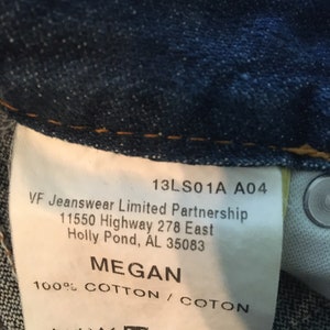 May include: A white tag with black text on a blue denim background. The tag says "13LS01A A04 VF Jeanswear Limited Partnership 11550 Highway 278 East Holly Pond, AL 35083 MEGAN 100% COTTON / COTON". There are also care instructions on the tag.