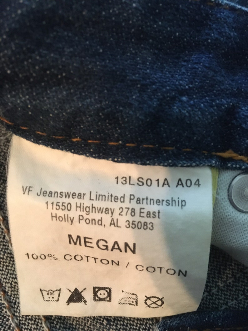 May include: A white tag with black text on a blue denim background. The tag says "13LS01A A04 VF Jeanswear Limited Partnership 11550 Highway 278 East Holly Pond, AL 35083 MEGAN 100% COTTON / COTON". There are also care instructions on the tag.