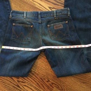 May include: A pair of blue denim jeans with a brown leather patch that says "Wrangler" on the back pocket. The jeans are laid flat on a wooden surface with a measuring tape.