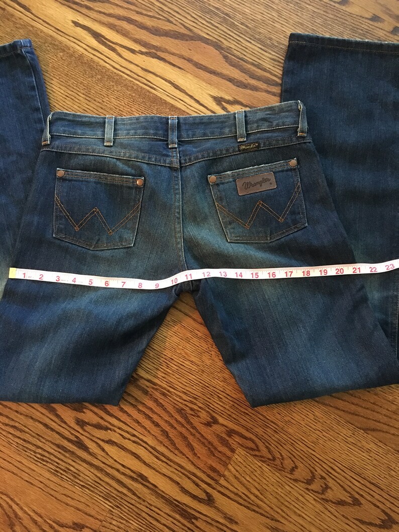 May include: A pair of blue denim jeans with a brown leather patch that says "Wrangler" on the back pocket. The jeans are laid flat on a wooden surface with a measuring tape.