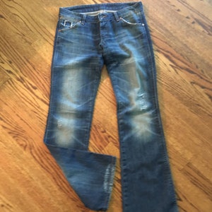 May include: A pair of blue denim jeans with faded and distressed details. The jeans have a bootcut style and are made of a soft, comfortable fabric.