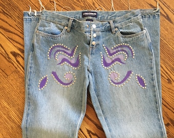 May include: Light blue denim jeans with a purple and silver design on the front. The jeans have a button fly and the brand name "LONDONJEAN" is printed on the back waistband.