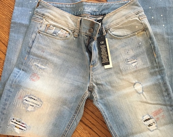 May include: Light blue distressed denim jeans with white paint splatters and decorative patches. The jeans have a 'VS Hipster' label and a size tag.