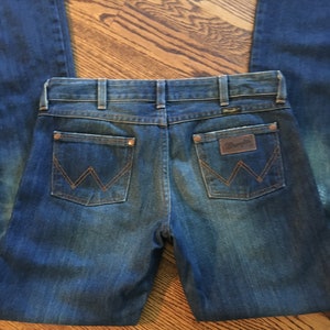 May include: A pair of blue denim jeans with a classic five-pocket design. The back pockets feature a stitched 'W' logo and the brand name 'Wrangler' is visible on the right back pocket.