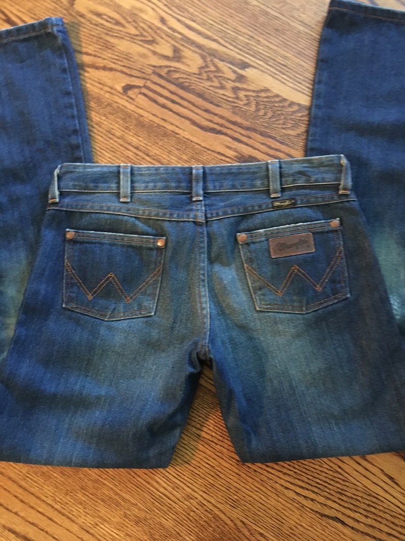 May include: A pair of blue denim jeans with a classic five-pocket design. The back pockets feature a stitched 'W' logo and the brand name 'Wrangler' is visible on the right back pocket.
