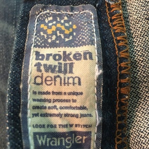 May include: A close-up of a Wrangler denim label with the text "broken twill denim" and a description of the fabric's unique weaving process. The label is beige with blue text and a small yellow graphic.