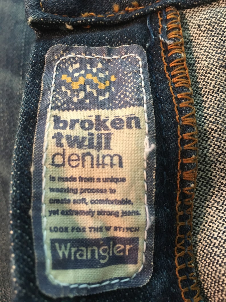 May include: A close-up of a Wrangler denim label with the text "broken twill denim" and a description of the fabric's unique weaving process. The label is beige with blue text and a small yellow graphic.