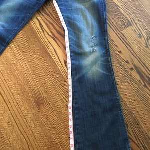 May include: A pair of blue denim jeans with a distressed look. The jeans are measured with a tape measure, showing a length of 32 inches.