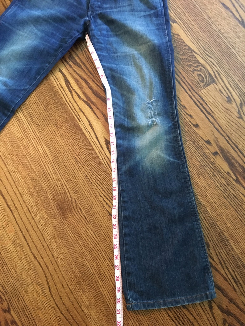 May include: A pair of blue denim jeans with a distressed look. The jeans are measured with a tape measure, showing a length of 32 inches.