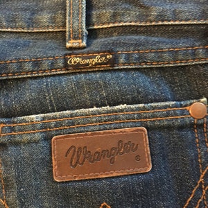May include: Close-up of a pair of blue denim jeans with a brown leather Wrangler label. The label has the Wrangler logo and a registered trademark symbol.