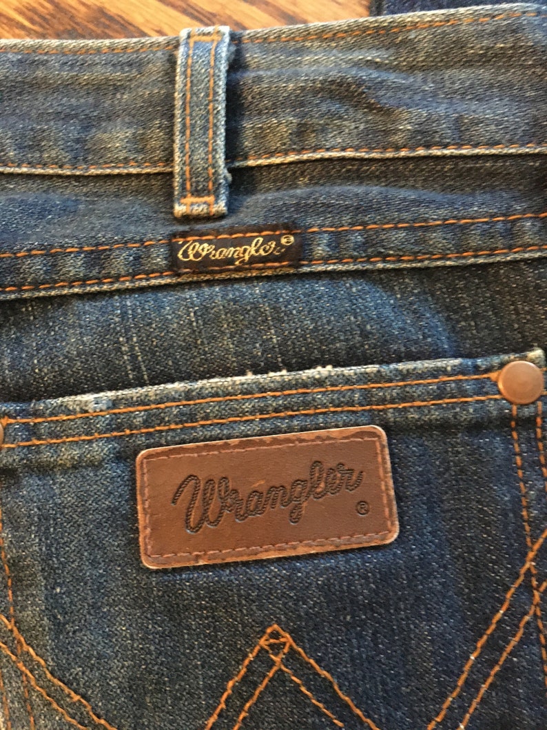 May include: Close-up of a pair of blue denim jeans with a brown leather Wrangler label. The label has the Wrangler logo and a registered trademark symbol.