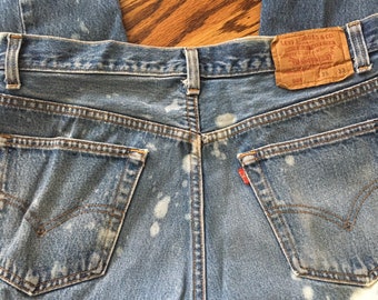 May include: A pair of blue denim jeans with white bleach splatters. The jeans have a brown leather patch with the text "Levi Strauss & Co.", "Quality Clothing", "501", "35", and "33".