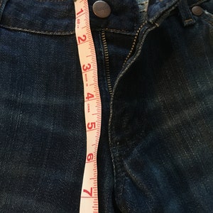 May include: A close-up of a pair of blue denim jeans with a zipper and a button closure. A measuring tape is placed along the side of the jeans, showing measurements in inches.