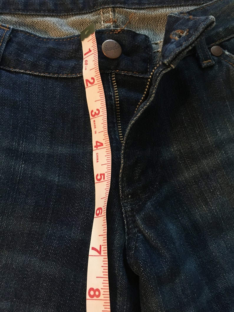 May include: A close-up of a pair of blue denim jeans with a zipper and a button closure. A measuring tape is placed along the side of the jeans, showing measurements in inches.
