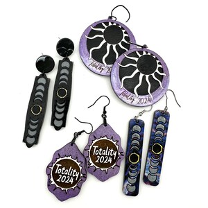 Puede incluir: Un conjunto de seis pendientes de madera con un esquema de color morado y negro. Los pendientes tienen forma de sol, luna creciente y rectángulo con un diseño de luna creciente. Los pendientes están decorados con el texto "Totality 2024".