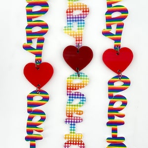 May include: Three rainbow-colored acrylic ornaments spelling out the word "PRIDE" with red heart accents. The ornaments are strung together with silver rings.