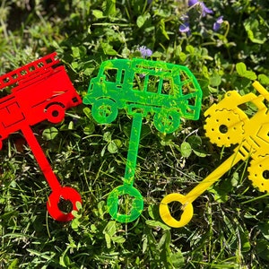 May include: Three colorful toy vehicles made of plastic. A red fire truck, a green van, and a yellow monster truck. Each vehicle is attached to a stick.