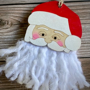 May include: A wooden Santa Claus ornament with a white yarn beard. The ornament has a red hat and a painted face with rosy cheeks.