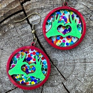 May include: A pair of round earrings with a red frame and a colorful polka dot background. The earrings feature a green hand-shaped design forming a heart. The earrings hang from gold-colored ear wires.