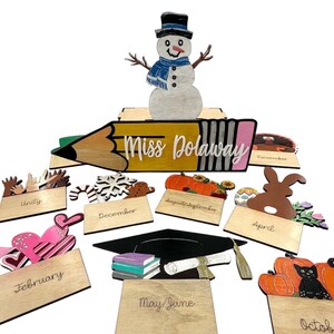 May include: Wooden classroom decor featuring a pencil, snowman, and monthly themed decorations. The pencil reads "Miss Dolaway" and each month has a different image. The months are January, February, March, April, May, June, July, August, September, October, November, and December.