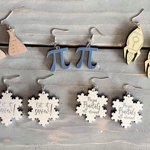 May include: A set of ten wooden earrings featuring various designs. The earrings are in the shape of snowflakes, beakers, rockets, and the mathematical symbol pi. Some of the earrings have text on them, including "Let it snow!" and "Frozen fractals all around".