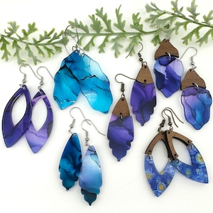 May include: A collection of various dangle earrings featuring blue and purple alcohol ink patterns on acrylic shapes, some accented with dark wood toppers and silver-toned metal hooks, displaying diverse geometric and organic silhouettes.