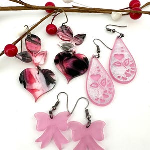 May include: A set of pink and white acrylic earrings. The earrings are in the shape of hearts, bows, and teardrops. The teardrop earrings have a white heart design.