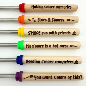 Puede incluir: Seis pinchos de madera con puntas metálicas y mangos de colores. Cada pincho tiene una frase diferente sobre s'mores grabada en la madera. Las frases son: "Making s'more memories", "Stars & Smores", "S'MORE fun with friends", "My s'more is a hot mess", "Needing s'more campfires", y "You want s'more of this?"