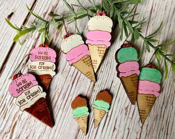 May include: Wooden earrings shaped like ice cream cones with different flavors. The earrings have a pink, white, and green ice cream design with a red cherry on top. The earrings are decorated with the text "We all scream for ice cream!"