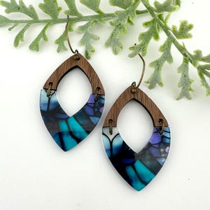 May include: A pair of teardrop earrings with a wooden top and a colorful, abstract design. The earrings feature a dark wood grain top with a clear, resin-like bottom. The bottom portion has a blue, teal, and purple pattern. The earrings hang from bronze-colored hooks.