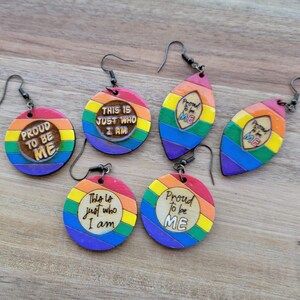 May include: Set of six rainbow-colored wooden earrings with different pride-themed messages. The earrings are round and oval shaped with a rainbow stripe pattern. The messages on the earrings include "Proud to be Me", "This is just who I am", and "Proud to be Me".