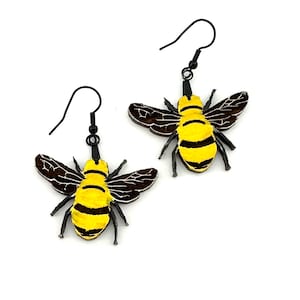 May include: A pair of dangle earrings featuring a yellow and black bee design. The bees are made of wood and have a glossy finish. The earrings are attached to black metal hooks.