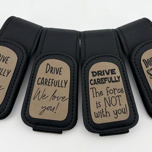 May include: Four black car visor clips with various phrases in white text on a tan background. The phrases include "Drive Carefully I love you!", "Drive Carefully We love you!", "Drive Carefully The force is NOT with you!", and "Don't do STUPID SHIT! - Your Wife".