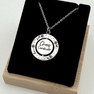 May include: A silver-colored pendant necklace with a round charm. The charm has the words "Chaos Coordinator" in a script font, surrounded by a floral design. The necklace is displayed in a black velvet box.